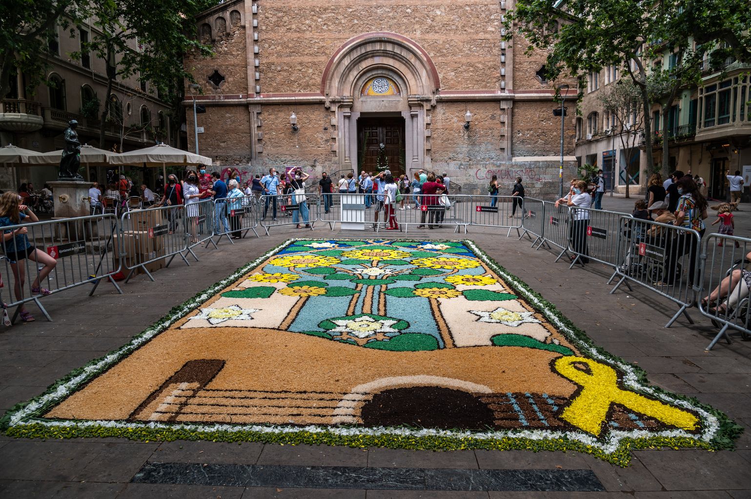 Vista de la gran catifa floral de la plaça de la Virreina creada amb motiu del Corpus. Al voltant, gent mirant i al fons, l'església de Sant Joan on hi ha una petita font amb l'ou com balla