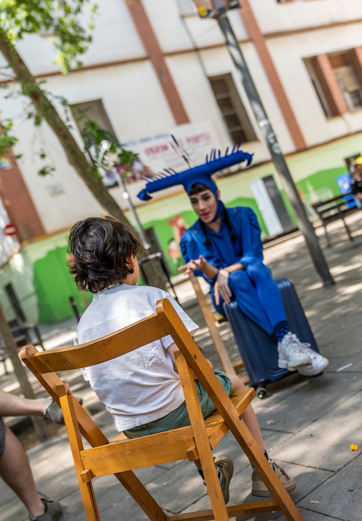 Un infant, assegut en una cadira, observa atentament les explicacions de Personaje Personaje durant l’activitat 'Colorete colorado' a la plaça del Sortidor.