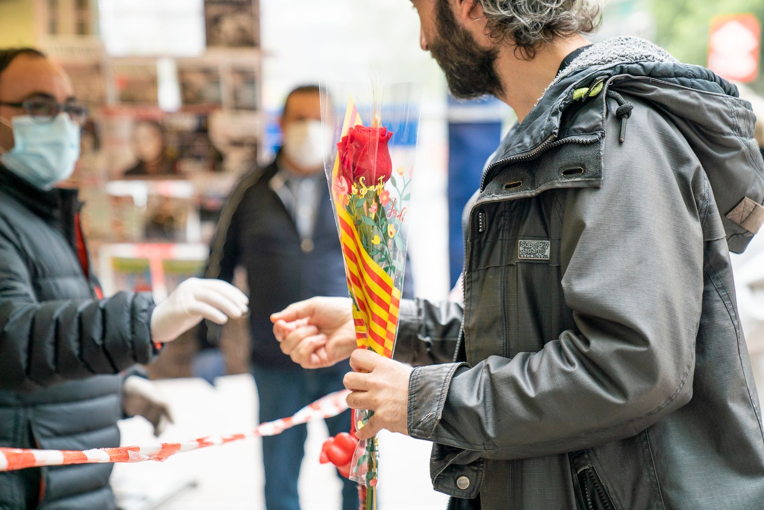 Un home rep el canvi per una rosa de mans del quiosquer de les distribuïdes per l’Ajuntament als quioscos de Diverscoop amb motiu de Sant Jordi. Es veu una cinta que precinta el voltant del quiosc per barrar el pas