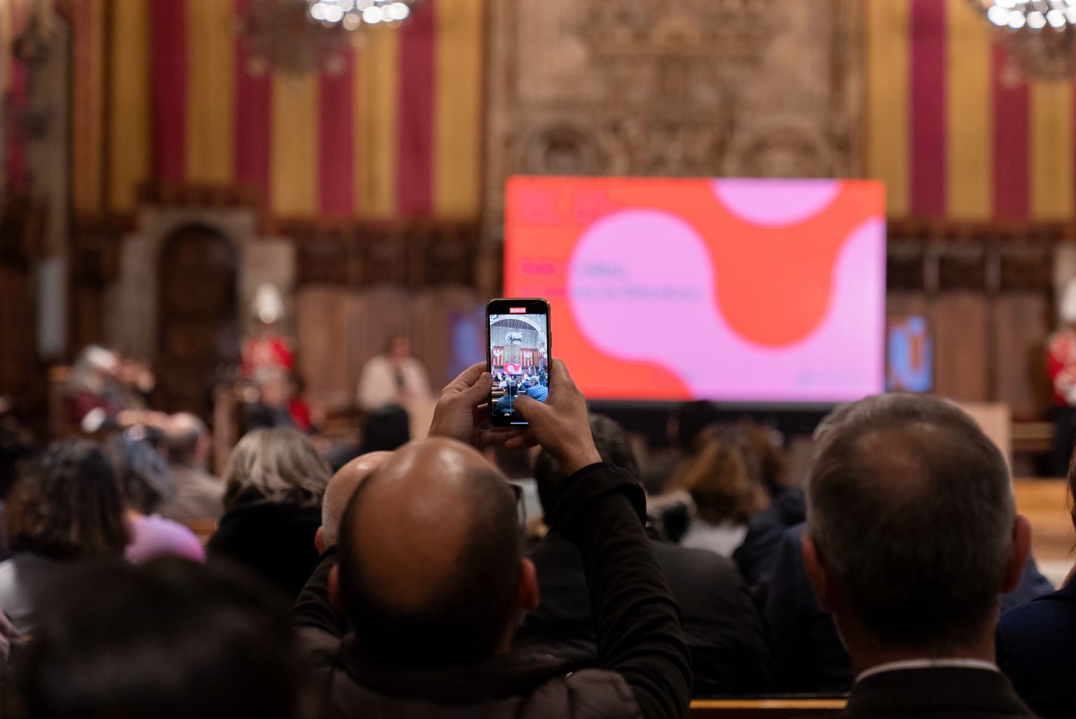 Un home del públic fa un vídeo amb el telèfon mòbil durant l’acte de presentació de la Biennal Ciutat i Ciència 2023, al Saló de Cent de l’Ajuntament de Barcelona.