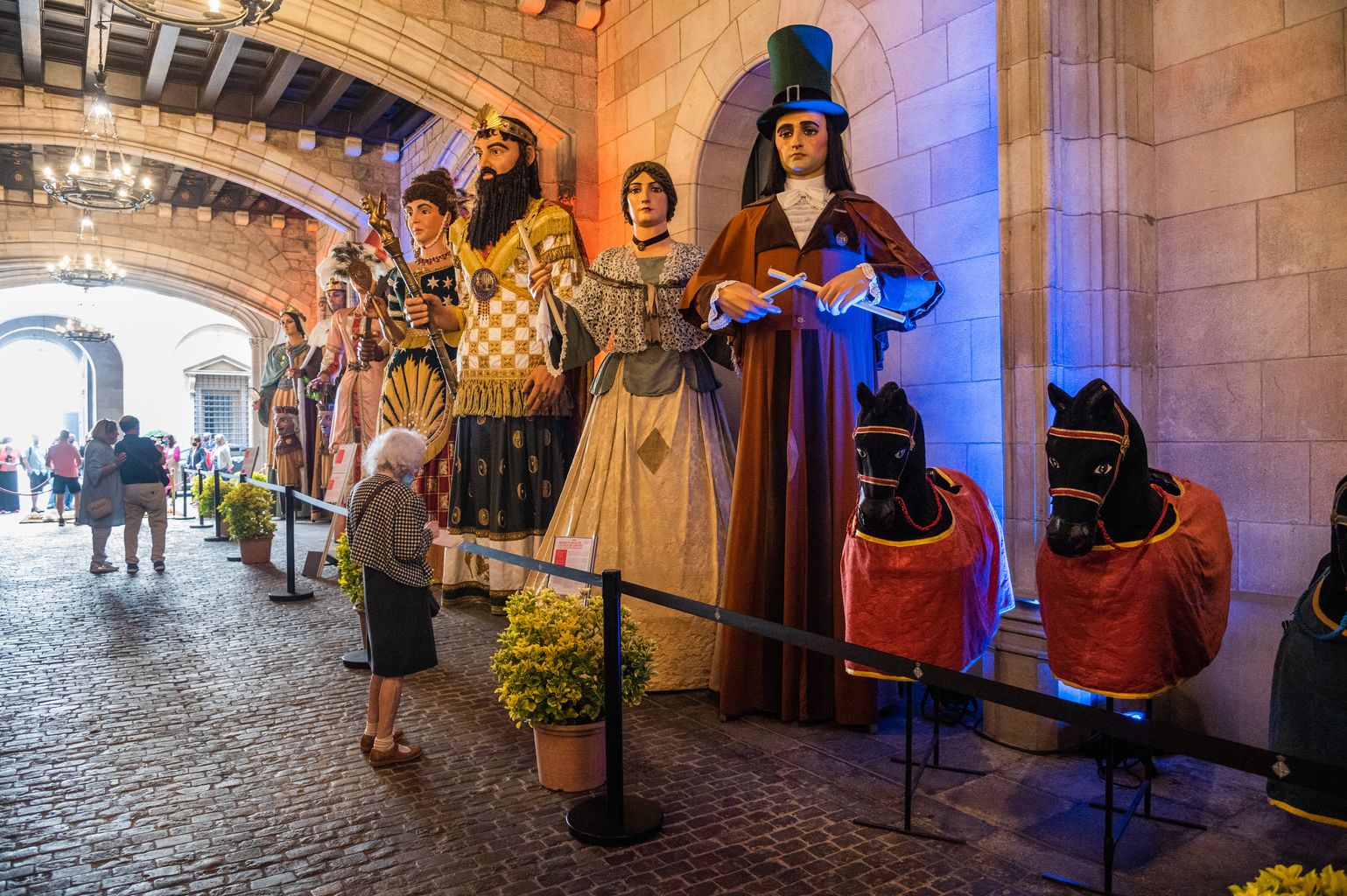 Gent visita l'exposició al pati de l'Ajuntament de Barcelona d'elements de la cultura popular de la ciutat. Una dona gran s'atura per mirar els Cavallets cotoners i els Gegants nous de la Casa de la Caritat o del Corpus