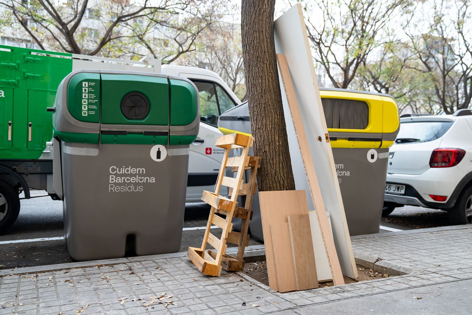 Puerta, estantes i palés de madera abandonados en un alcorque al lado de unos contenedores de Cuidem Barcelona Residus