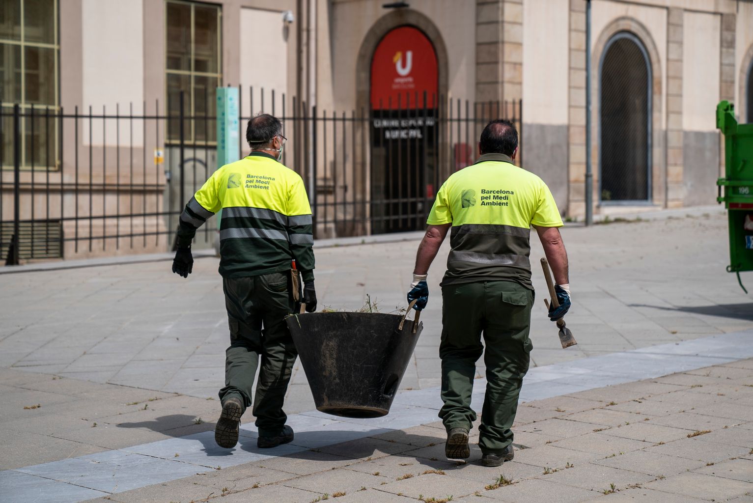 Dos jardiners transporten un cistell de males herbes mentre un agafa un rasclet i l'altre, amb una aixada, es dirigeix al camió