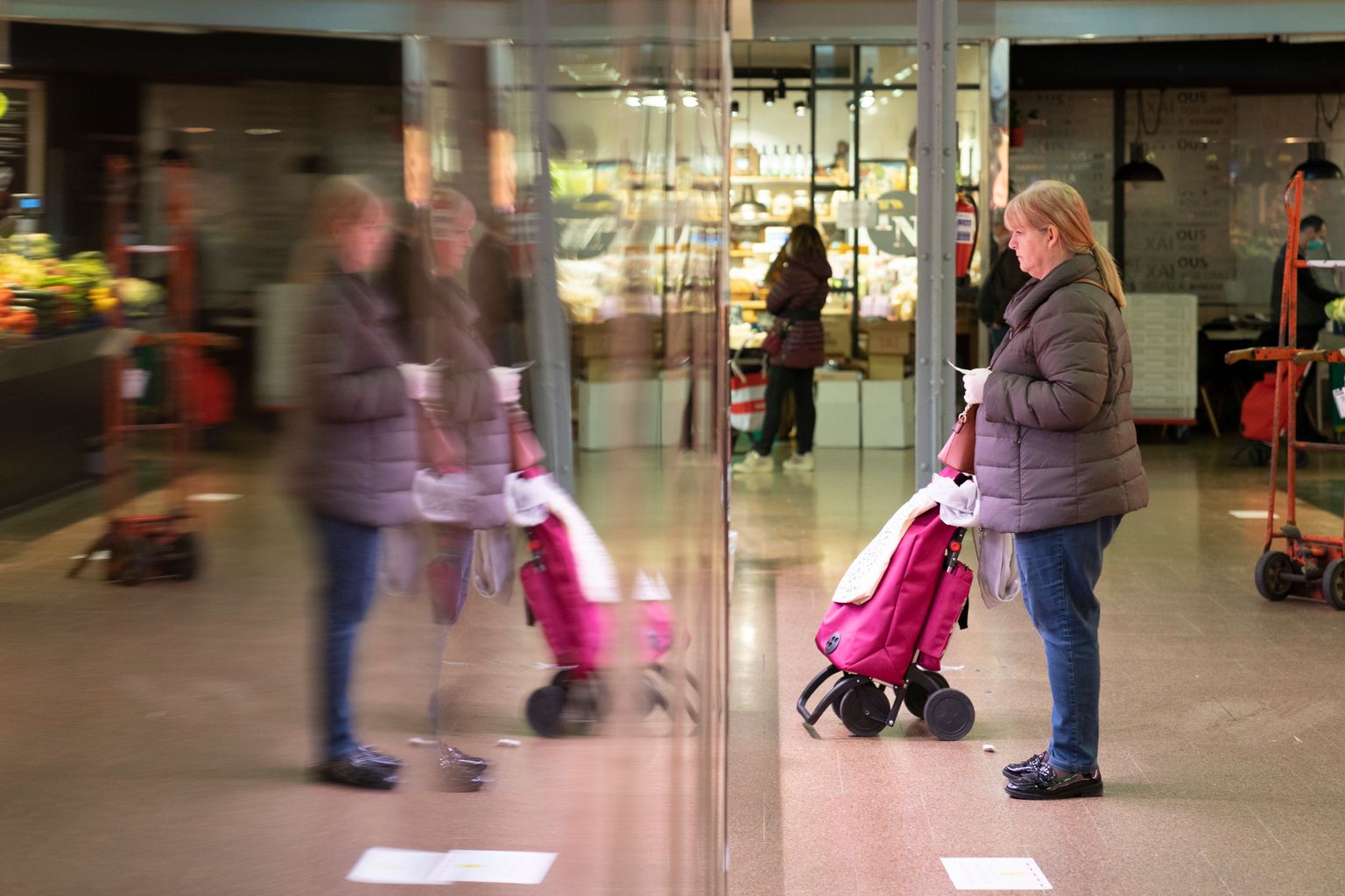 Mercat del Ninot. Clienta amb carretó d’anar a comprar, amb guants i un paper a la mà davant d’una parada mantenint la distància de seguretat establerta