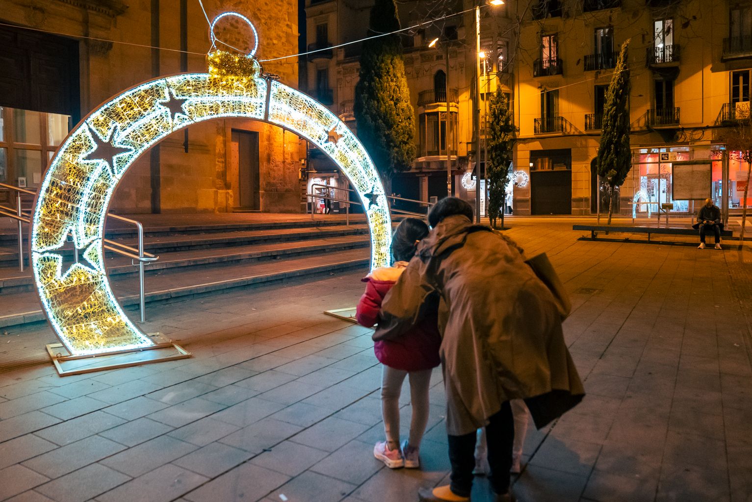 Una mujer con dos niños observan un tótem de luz navideña en forma de bola, iluminado ante la parroquia de Sant Vicenç de Sarrià, en la plaza de Sarrià