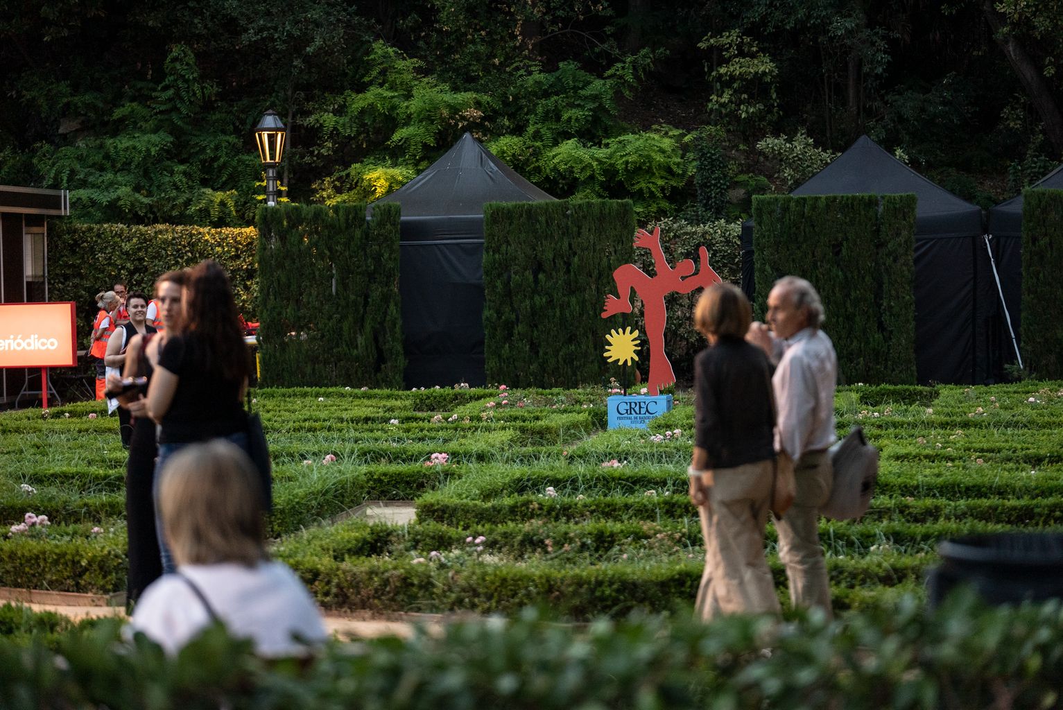 Persones caminant pels jardins del teatre Grec. Al fons, una escultura de promoció del festival Grec 23.
