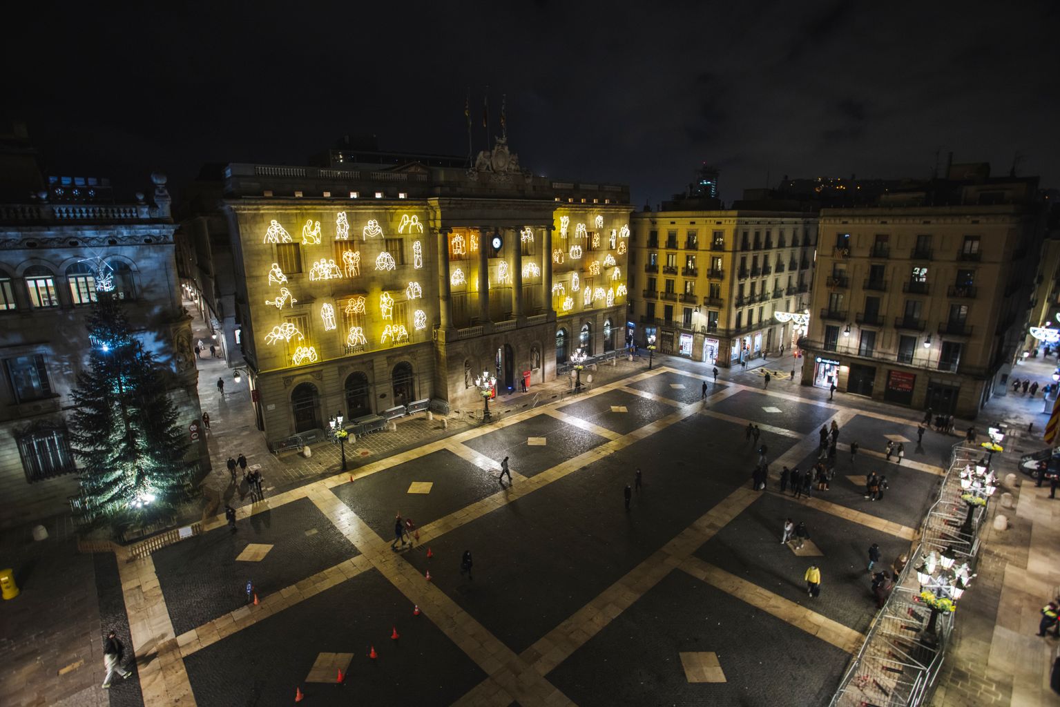 Vista de la plaça de Sant Jaume amb la façana de l'Ajuntament il·luminada amb escenes quotidianes de Ciutat Vella, una obra del col·lectiu RavalKm#0 amb motiu de les festes de Nadal. Al costat, es veu l'arbre de Nadal