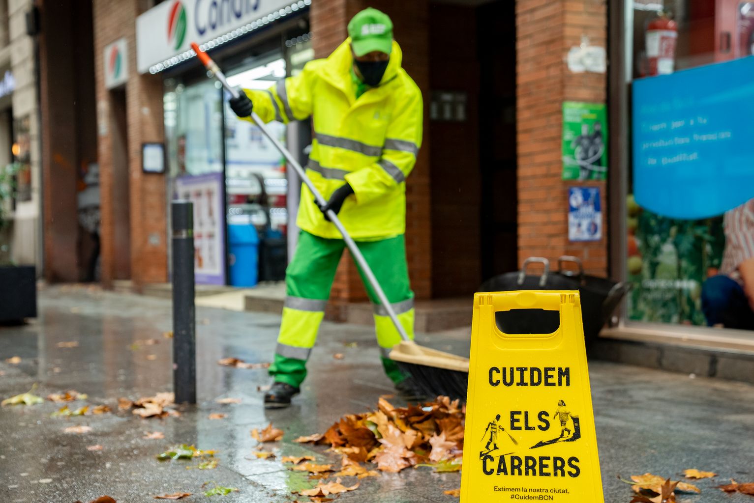 Un trabajador del Servicio de Limpieza barre las hojas secas de una acera de la rambla del Poblenou. Ha puesto un indicador de aviso con el letrero “Cuidem els carrers”