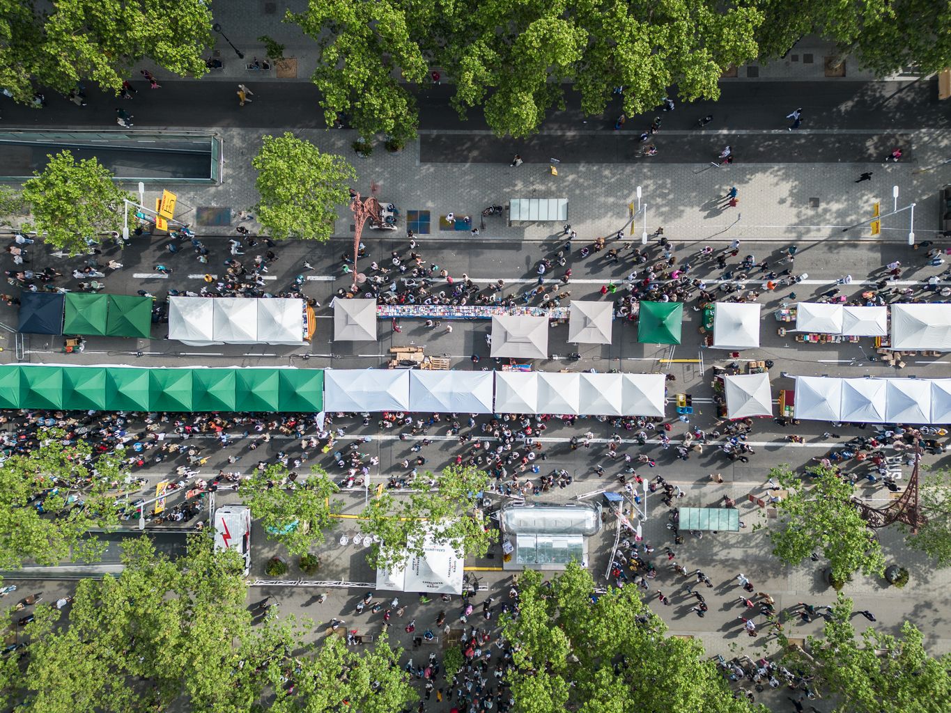 Vista aèria zenital de les parades de Sant Jordi instal·lades al passeig de Gràcia, a prop de l'entrada d'un aparcament subterrani i amb gent passejant entre les parades de roses i llibres. Es veu també la carpa de Catalunya Ràdio.