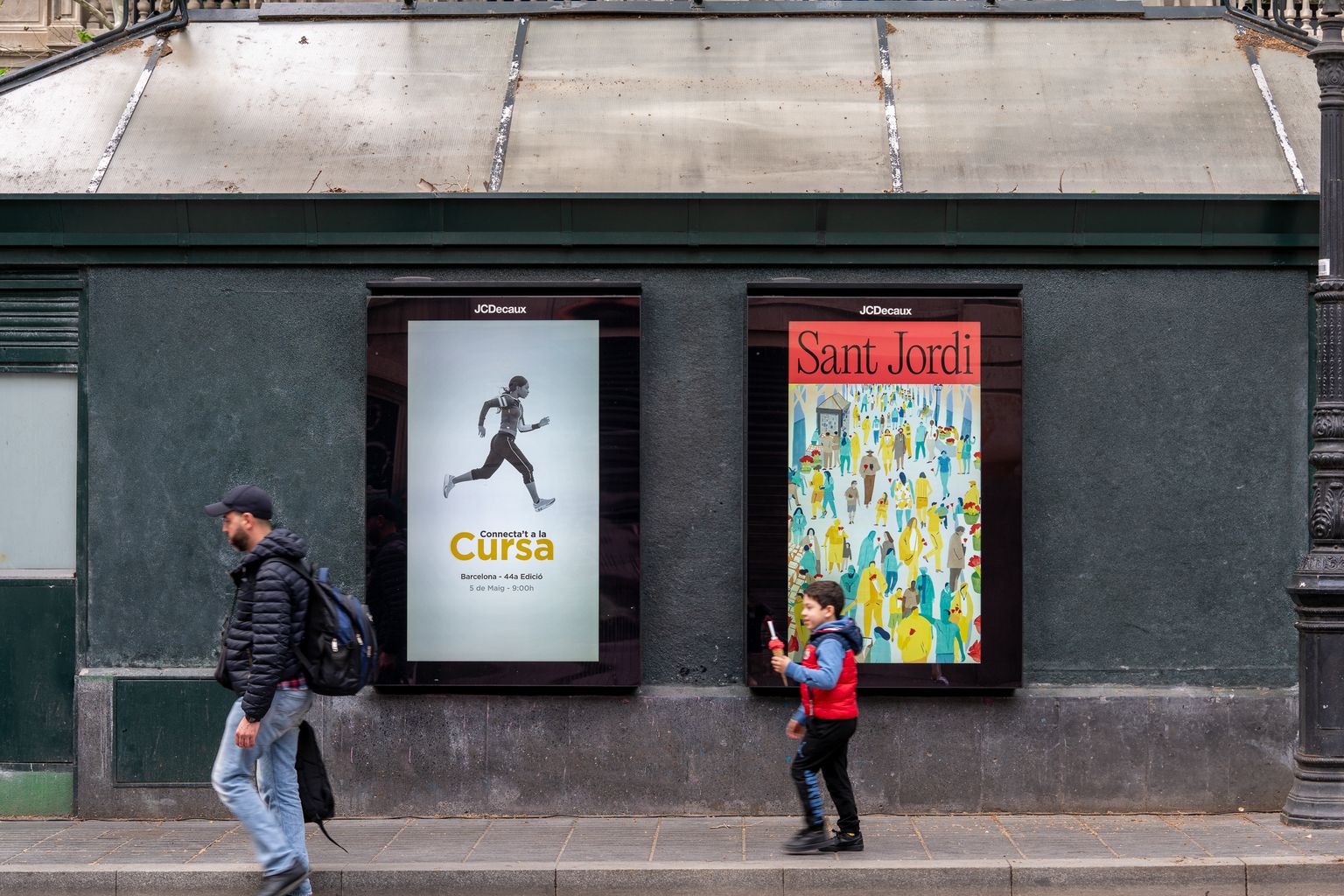 Personas caminando delante de un opi digital con la imagen gráfica de la campaña publicitaria de Sant Jordi 2024 en un quiosco de La Rambla.