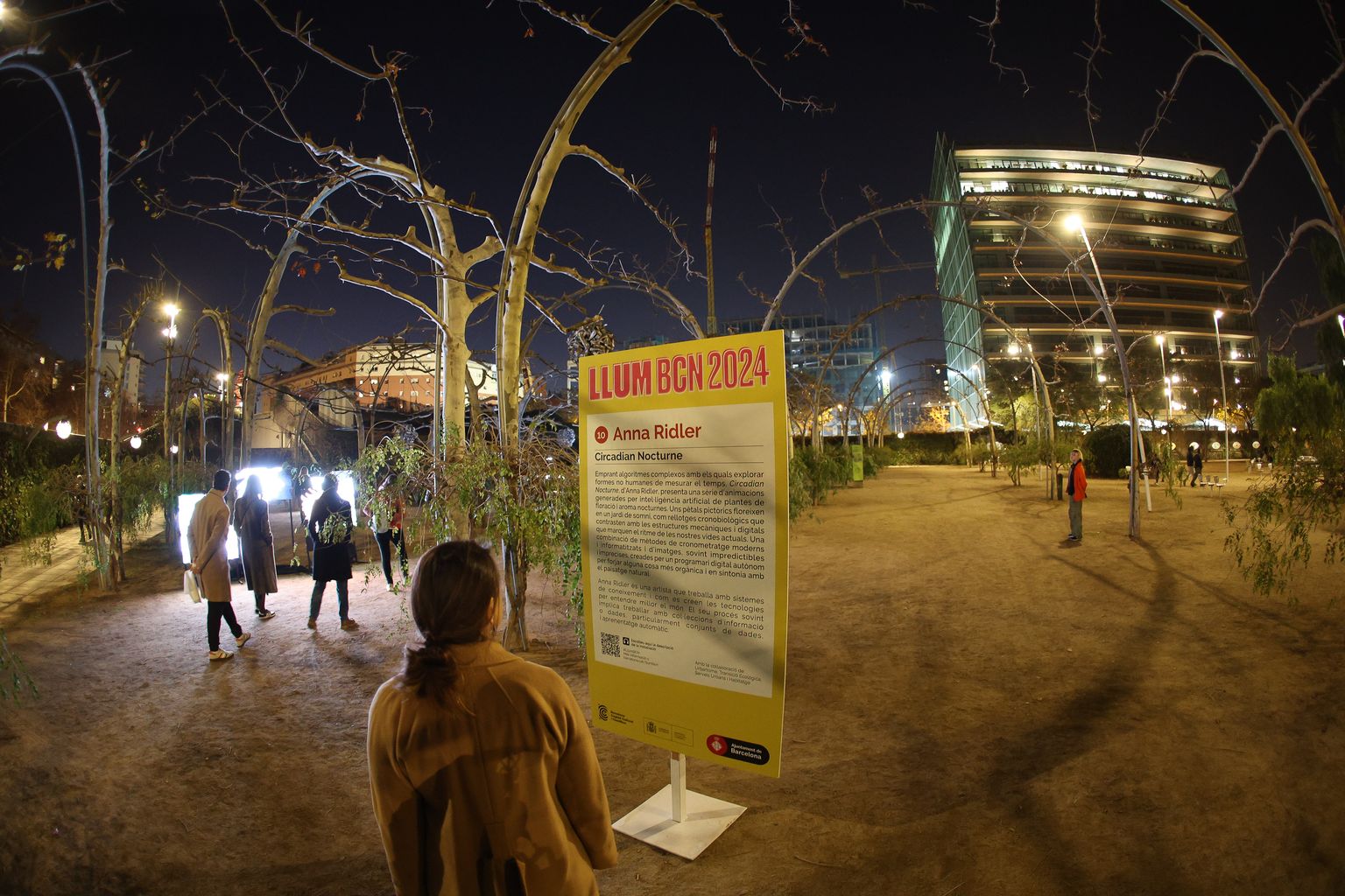 Persones passegen pel parc del Centre del Poblenou. Una dona mira el cartell del Llum BCN 2024. “Circadian Nocturne”, obra per Anna Ridler. Al Parc del Centre del Poblenou (Espai Voltes) - av. Diagonal, 130