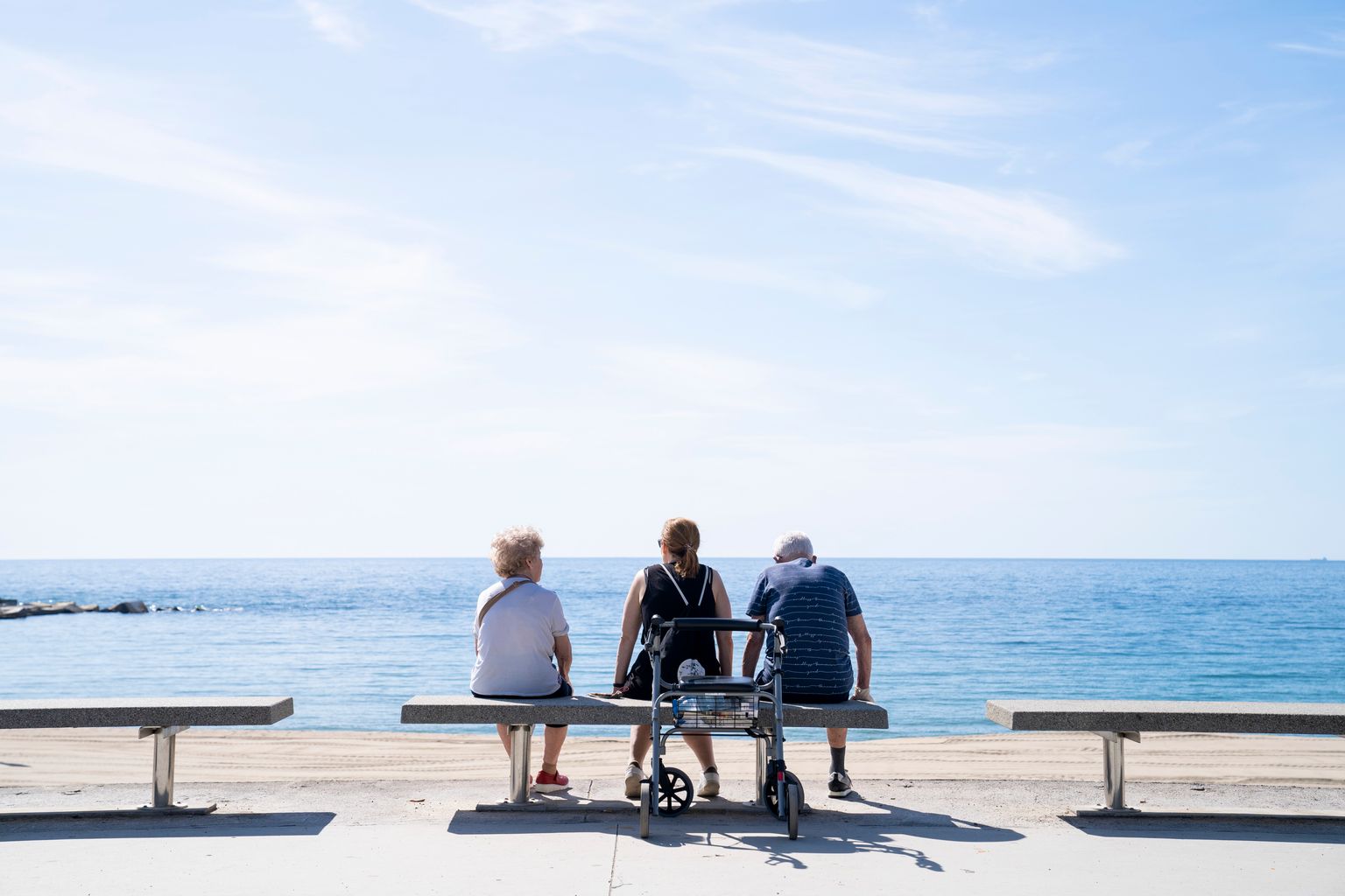 Dues persones grans amb una acompanyant estan assegudes als bancs del passeig Marítim del Bogatell. Al darrere, un caminador