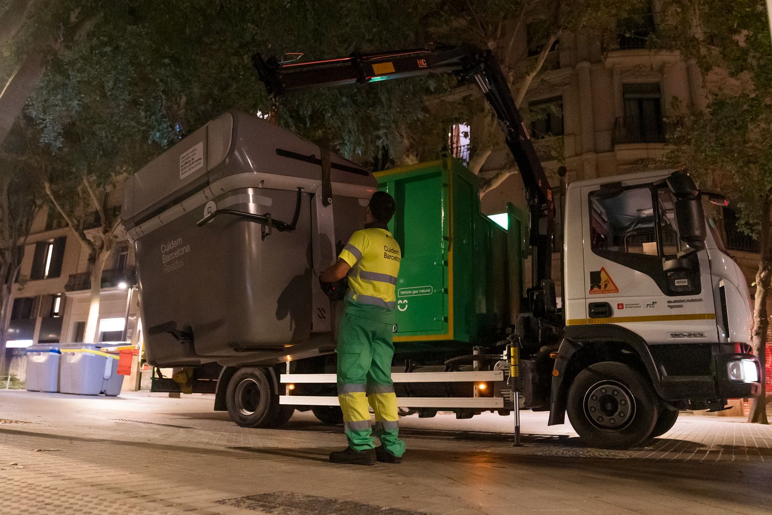 Un camión de Cuidem Barcelona cogiendo un contenedor de reciclaje con la grúa mientras un operario lo controla desde abajo.