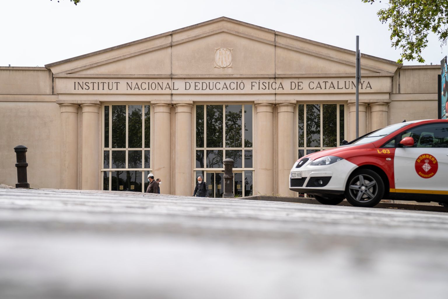 Treballadors entrant i sortint de l’Institut Nacional d’Educació Física de Catalunya. Davant hi ha aparcat un vehicle dels Bombers de Barcelona