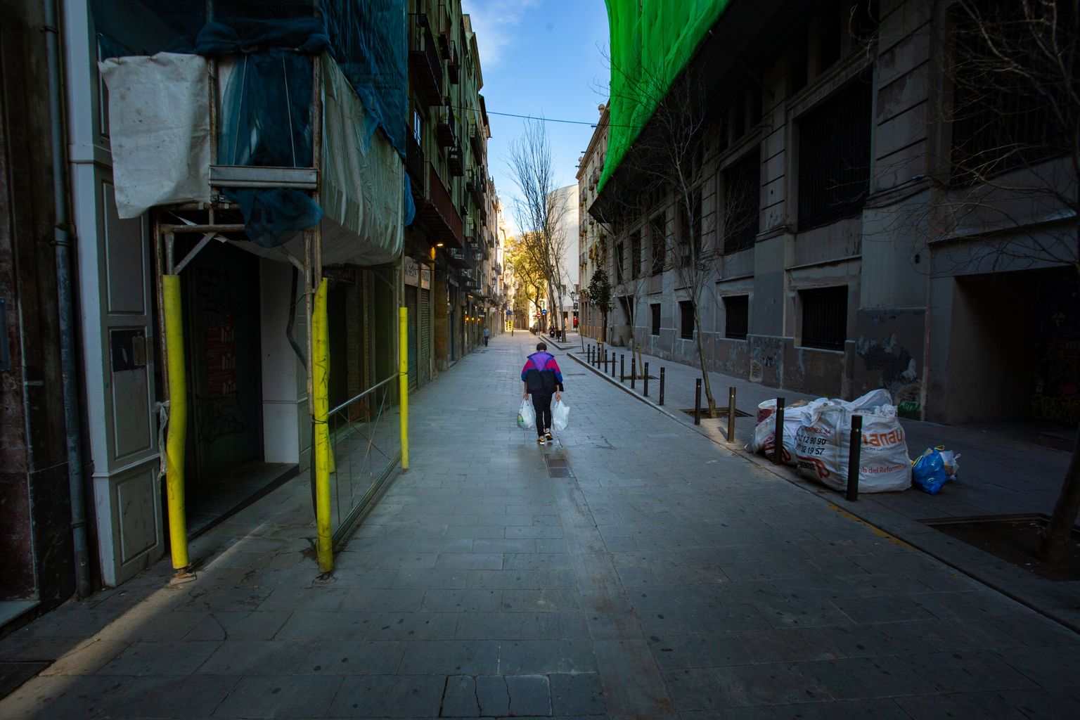 Carrer de l’Argenteria sense activitat ni turistes, un home camina carregat de bosses. Es veuen sacs de runes pendents de recollir davant d’una obra. Districte de Ciutat Vella. Barri de Sant Pere, Santa Caterina i la Ribera.