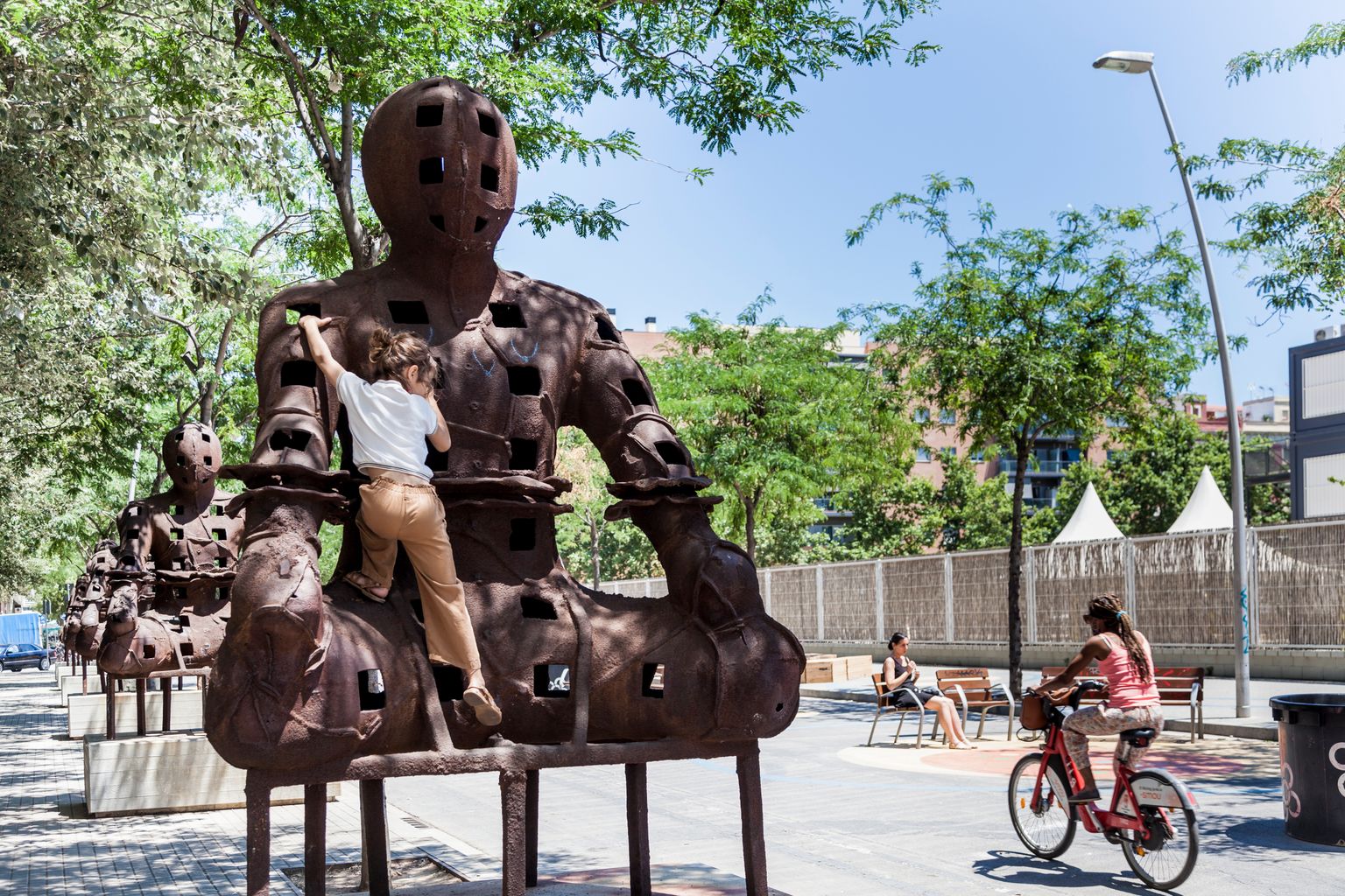 Una nena s’enfila per una de les escultures ‘Guardians’, a la superilla del Poblenou. Pel costat, al carrer de Sancho de Ávila, passa una ciclista