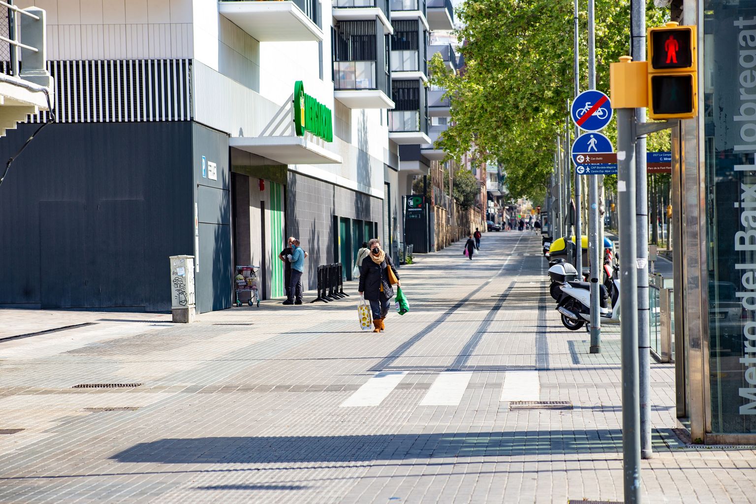 Activitat de persones al costat d’un supermercat amb gent caminant amb bosses de la compra a la gran via de les Corts Catalanes al voltant de Magòria. Es veu la parada de metro del Baix Llobregat. Districte de Sants-Montjuïc. Barri de la Bordeta.