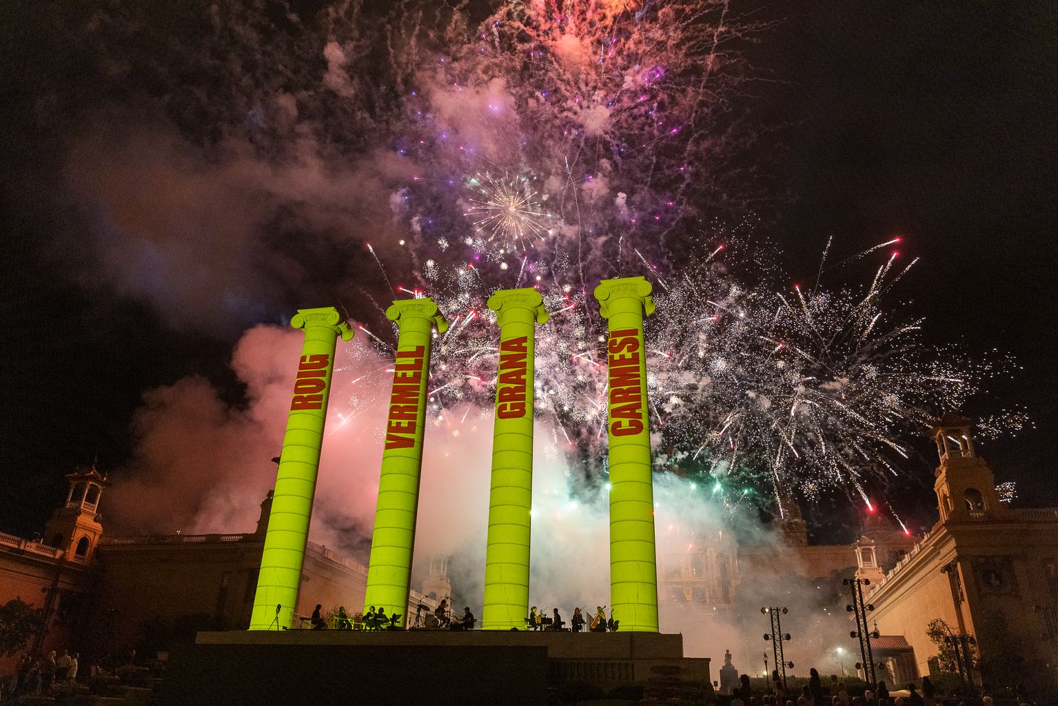 Les Quatre Columnes de Montjuïc il·luminades de groc amb les paraules “Roig, Vermell, Grana, Carmesí” de color vermell. Al fons, focs artificials.