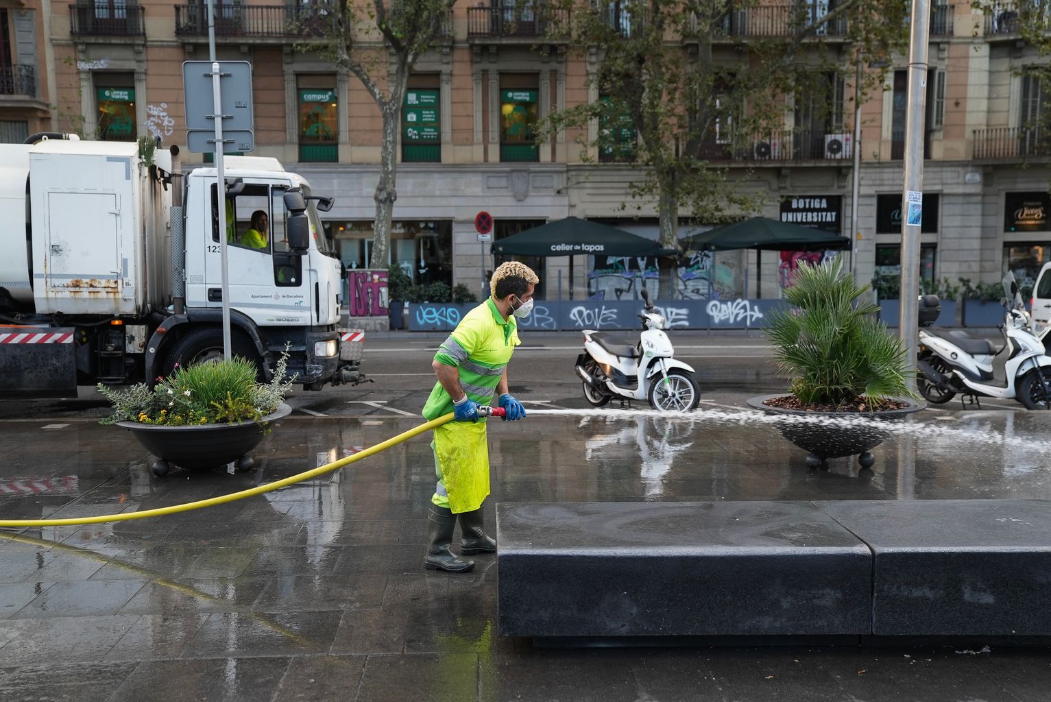 Un trabajador limpia con agua a presión el pavimento y un banco de piedra de la plaza de la Universitat