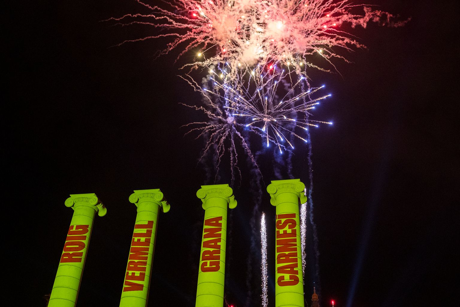 Les Quatre Columnes de Montjuïc il·luminades de groc amb les paraules “Roig, Vermell, Grana, Carmesí” de color vermell. Al fons, focs artificials.