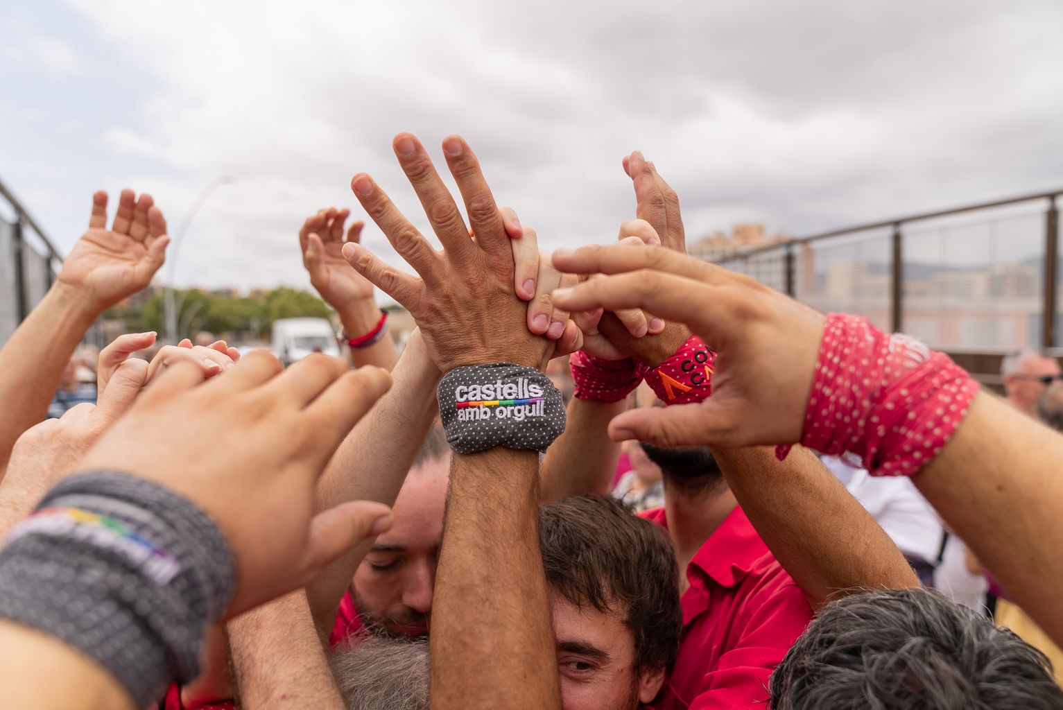 Detall de les mans fent pinya en un castell dels Castellers de Barcelona. Al centre, un mocador en un canell on llegim "Castells amb orgull"