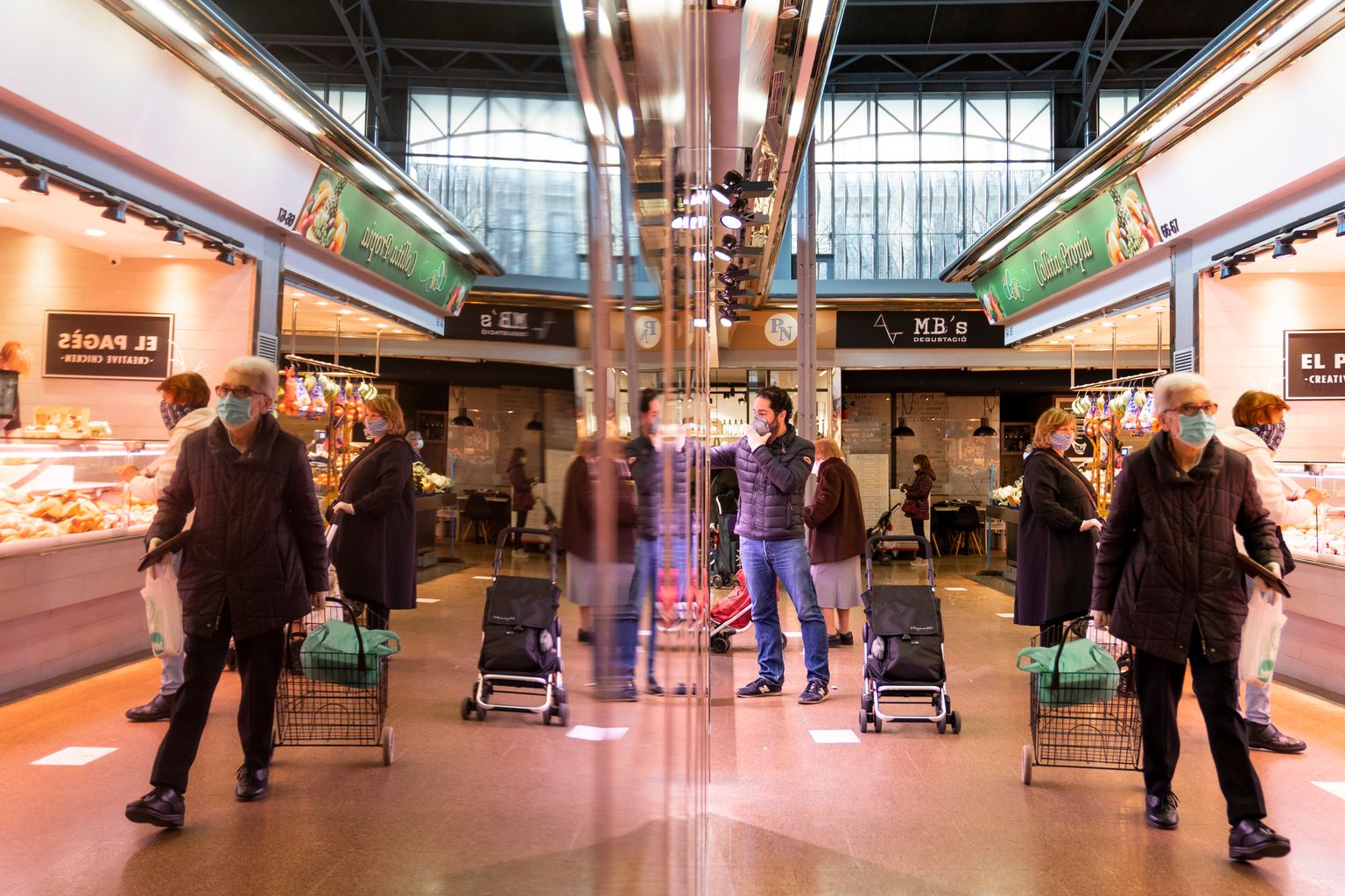 Mercat del Ninot. Gent amb carretons i mascaretes comprant a les diferents parades i mantenint la distància de seguretat entre ells. Imatge duplicada pel reflex en una superfície metàl·lica