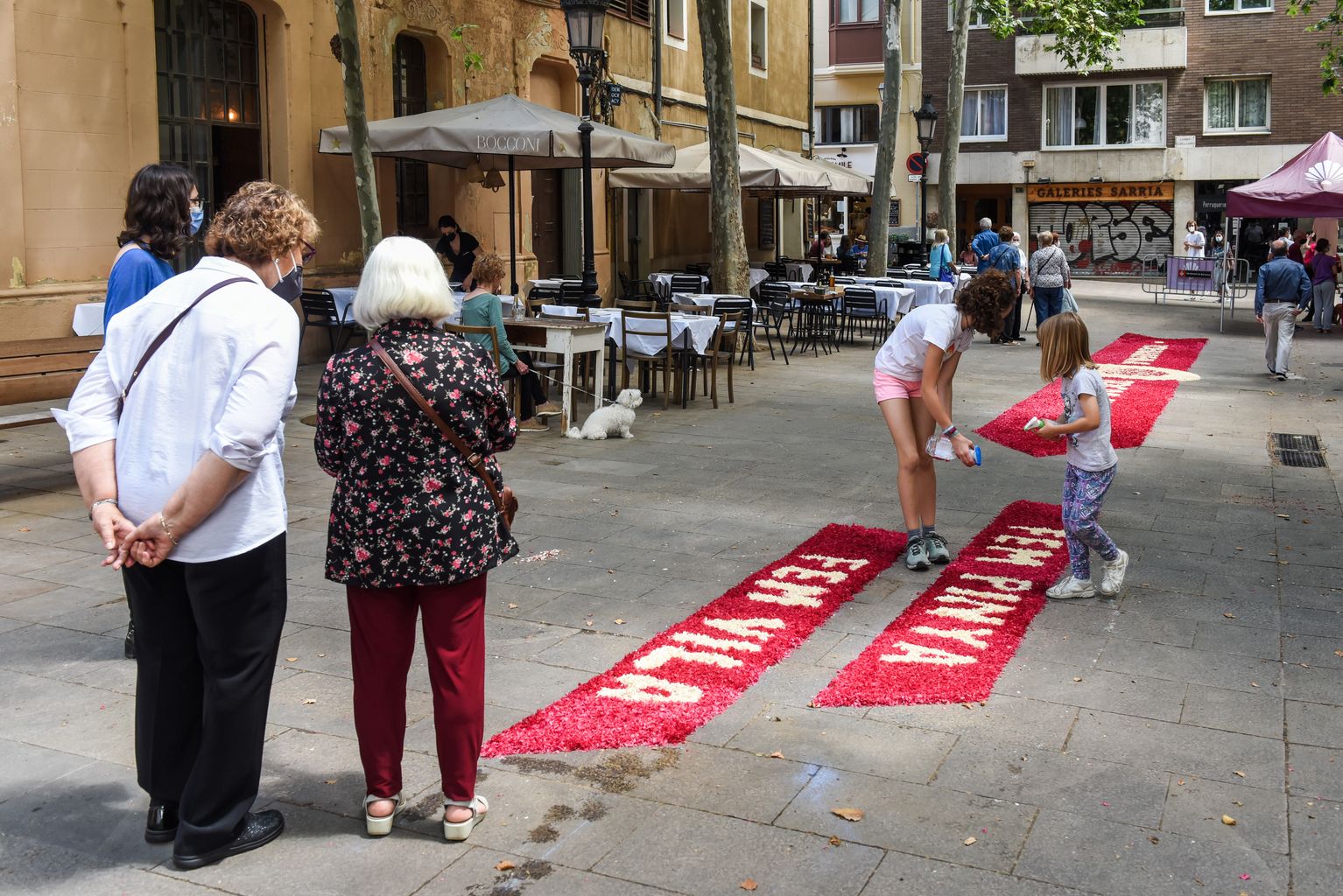 Nenes treballen en la creació de la catifa floral que fan els Castellers de Sarrià amb motiu de la festa del Corpus a la plaça del Consell de la Vila. Unes dones grans miren com les fan