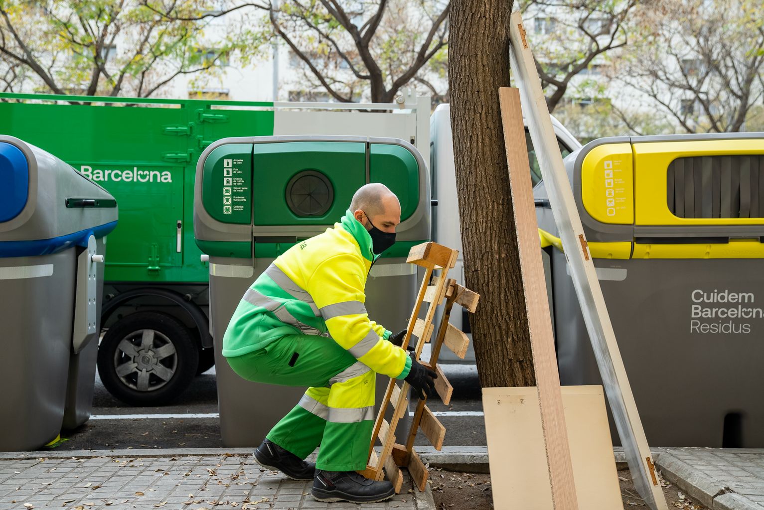 Un trabajador del servicio de limpieza (Cuidem Barcelona Residus) recoge unas maderas y muebles abandonados en el alcorque de un árbol junto a unos contenedores de reciclaje