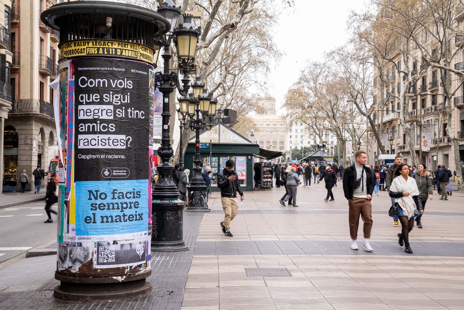 Tòtem a les Rambles amb el cartell de la campanya Barcelona antiracista que porta el lema "Com vols que sigui negre si tinc amics racistes?" i gent passejant pel carrer
