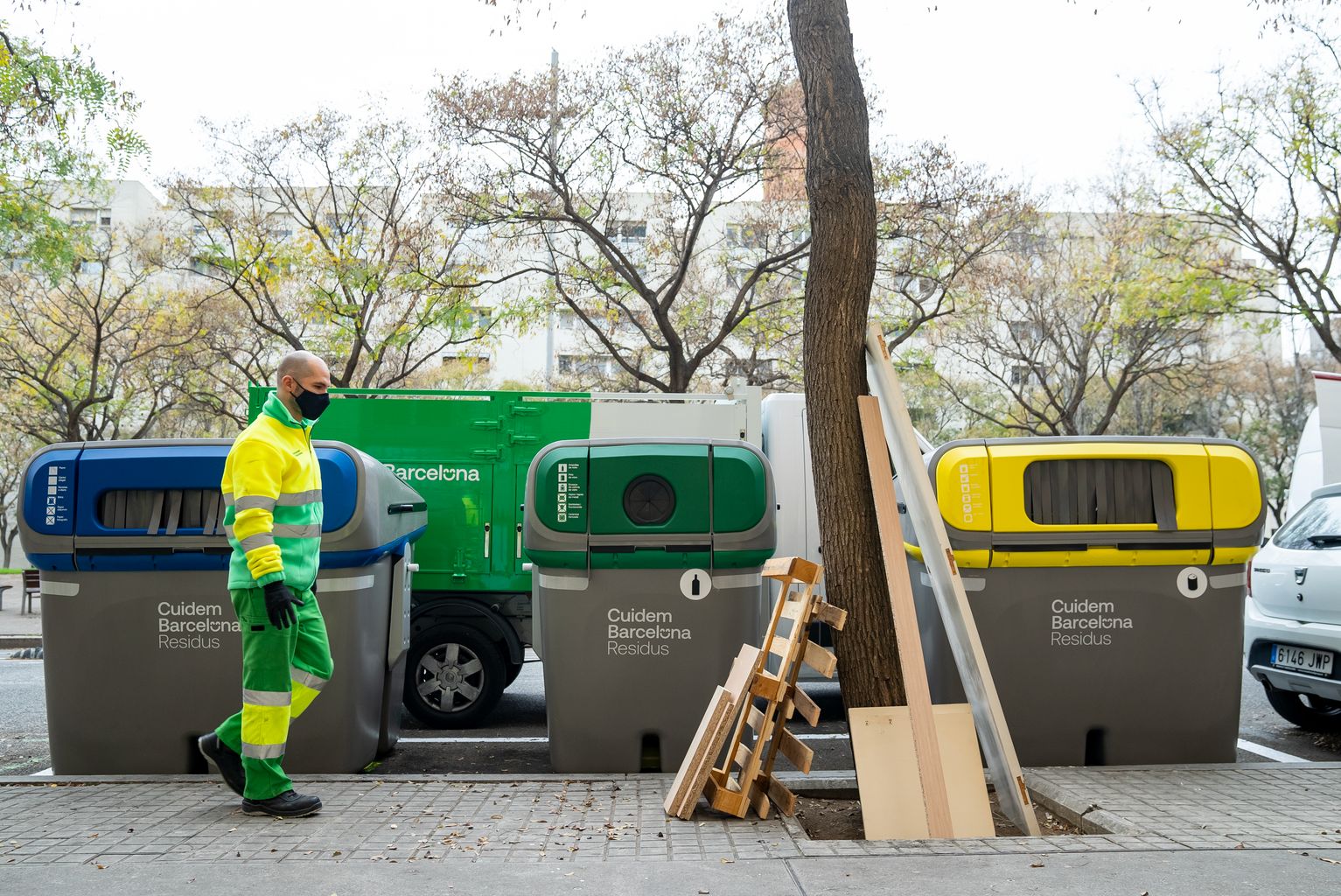 Un treballador del servei de neteja (Cuidem Barcelona Residus) s'apropa per a recollir unes fustes i mobles abandonats a l'escocell d'un arbre al costat d'uns contenidors de reciclatge