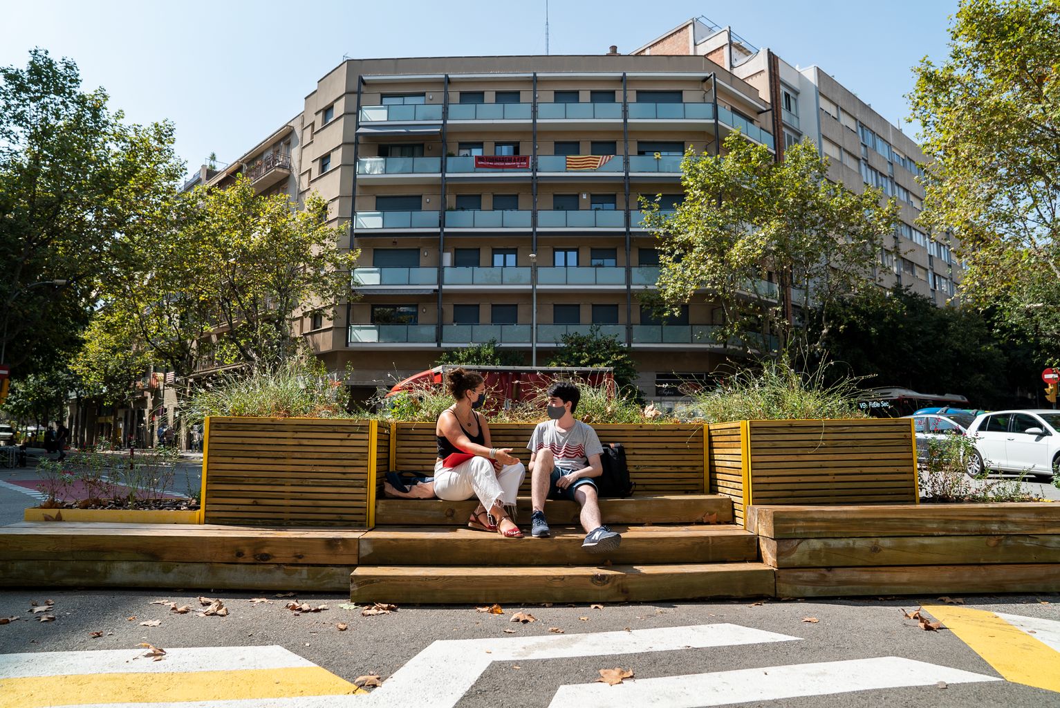 Dos joves de l'Institut Viladomat conversen asseguts a les jardineres amb bancs instal·lades a la zona pacificada al voltant del centre educatiu. Es veu la zona de la vorera ampliada pintada amb ratlles obliqües blanques i grogues