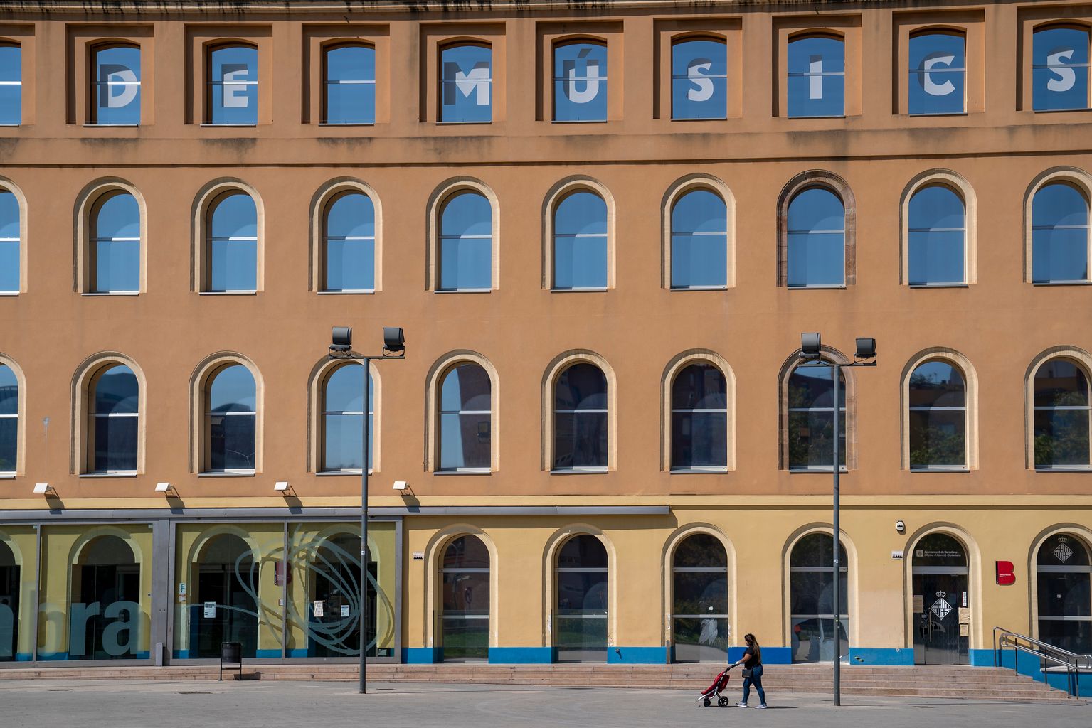 Plaça de Can Fabra amb una persona travessant-la amb un carret d’anar a comprar. Al darrere, l’edifici de Can Fabra que roman tancat