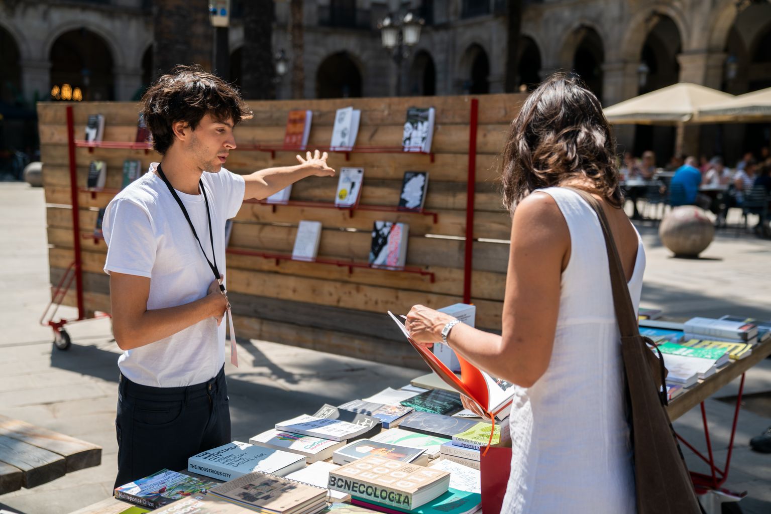 Un jove informa una visitant de la selecció de llibres sobre arquitectura que s’exposen als taulells a la plaça Reial