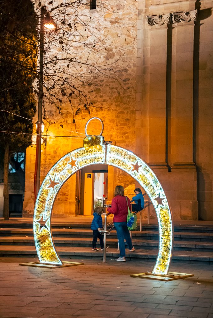 Dos niños y su madre juegan en las escaleras de la plaza de Sarrià, tras un tótem de luz navideña en forma de bola. Detrás, la parroquia de Sant Vicenç de Sarrià