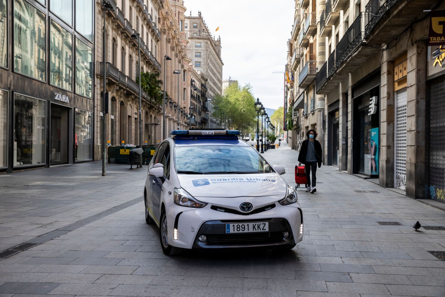 Avinguda del Portal de l’Àngel amb un cotxe de la Guàrdia Urbana i una persona amb mascareta caminant i portant un carretó d’anar a comprar