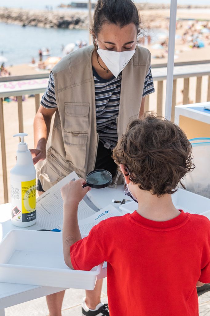 Meritxell Muray, responsable de l’activitat “Descobrim els microplàstics!”, mostra a un infant que participa al taller el contingut de la sorra de la platja