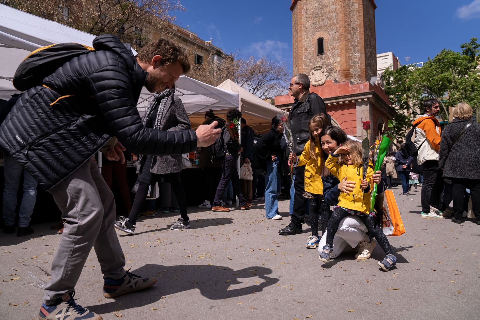 Un hombre fotografiando a su familia, que llevan unas rosas, entre los puestos de Sant Jordi de la plaza de la Vila de Gràcia