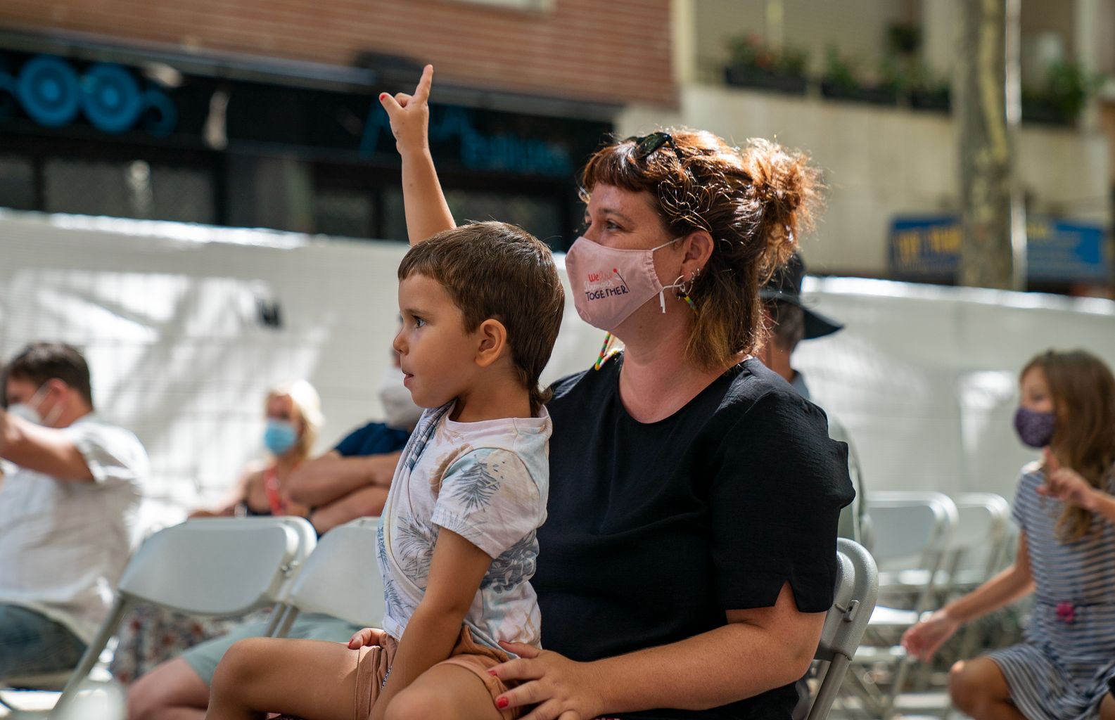 Un nen del públic assegut a la falda de la seva mare aixeca la mà per fer una pregunta durant el contacontes "Amunt i avall: pagesos, cucs i alls", una de les activitats de la Festa Major de Gràcia