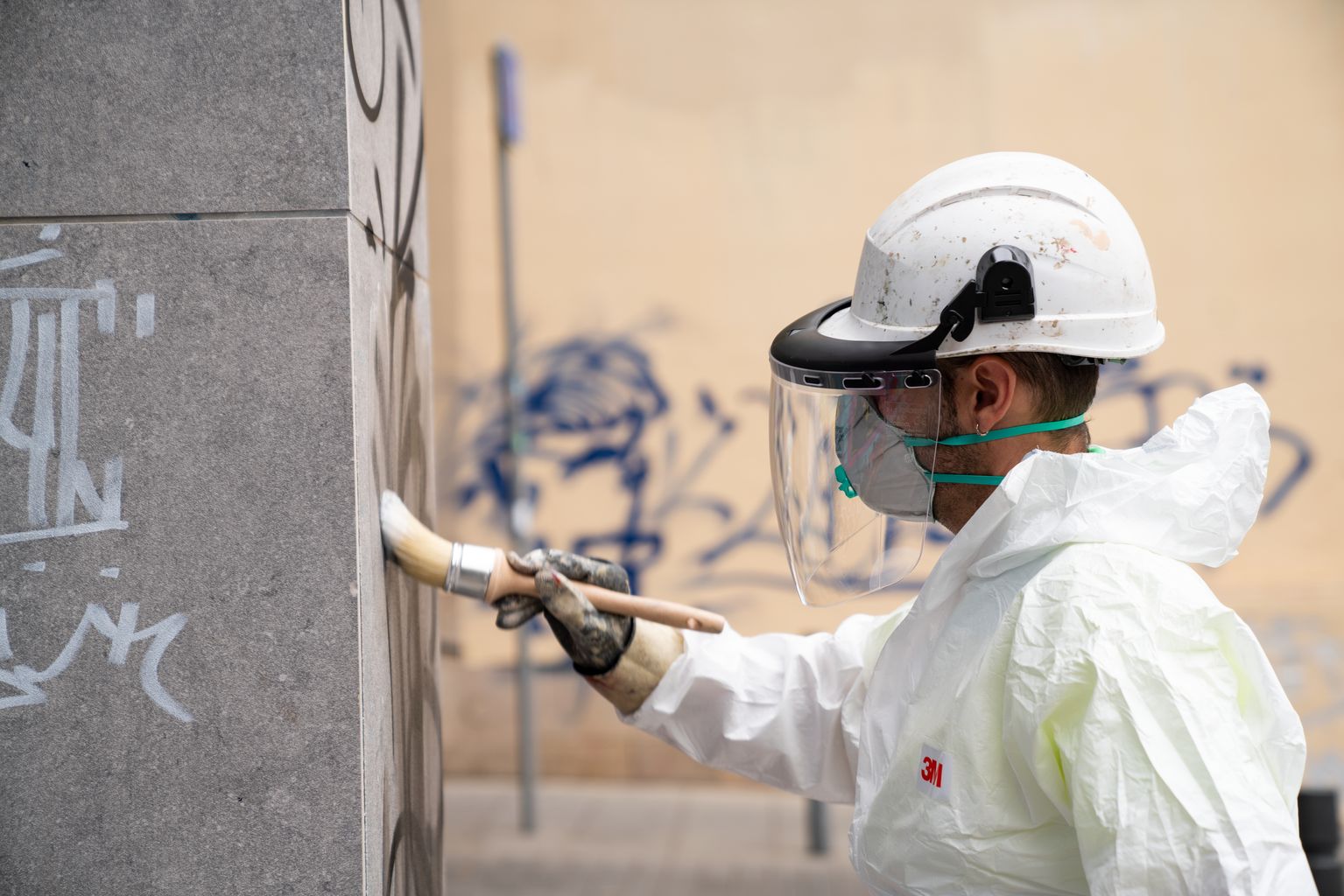 Un trabajador con el mono de protección y una mascarilla de seguridad aplica un producto sobre unos grafitis pintados en una pared para retirarlos