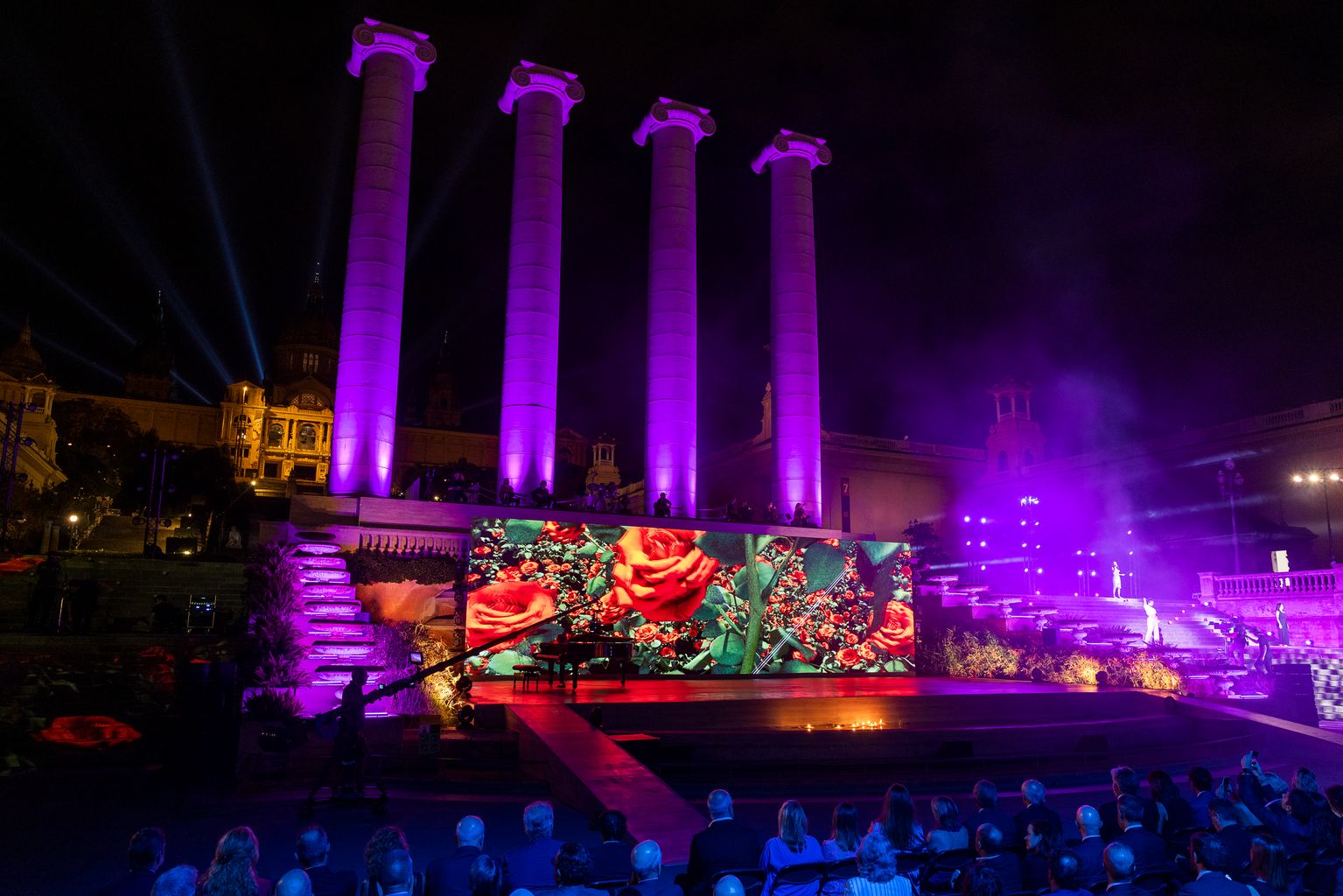 Pla general de l’acte institucional de la Diada. A l’escenari, un piano i una pantalla on apareix un estampat de roses. Al darrere, les Quatre Columnes de Montjuïc il·luminades.