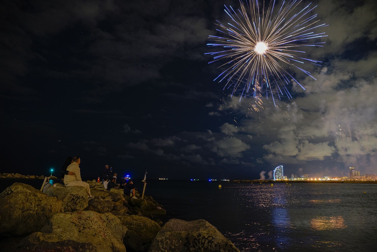 Gent asseguda a l'espigó seguint el 25è Festival Pirotècnic Internacional a la Platja de l'Espigó del Gas