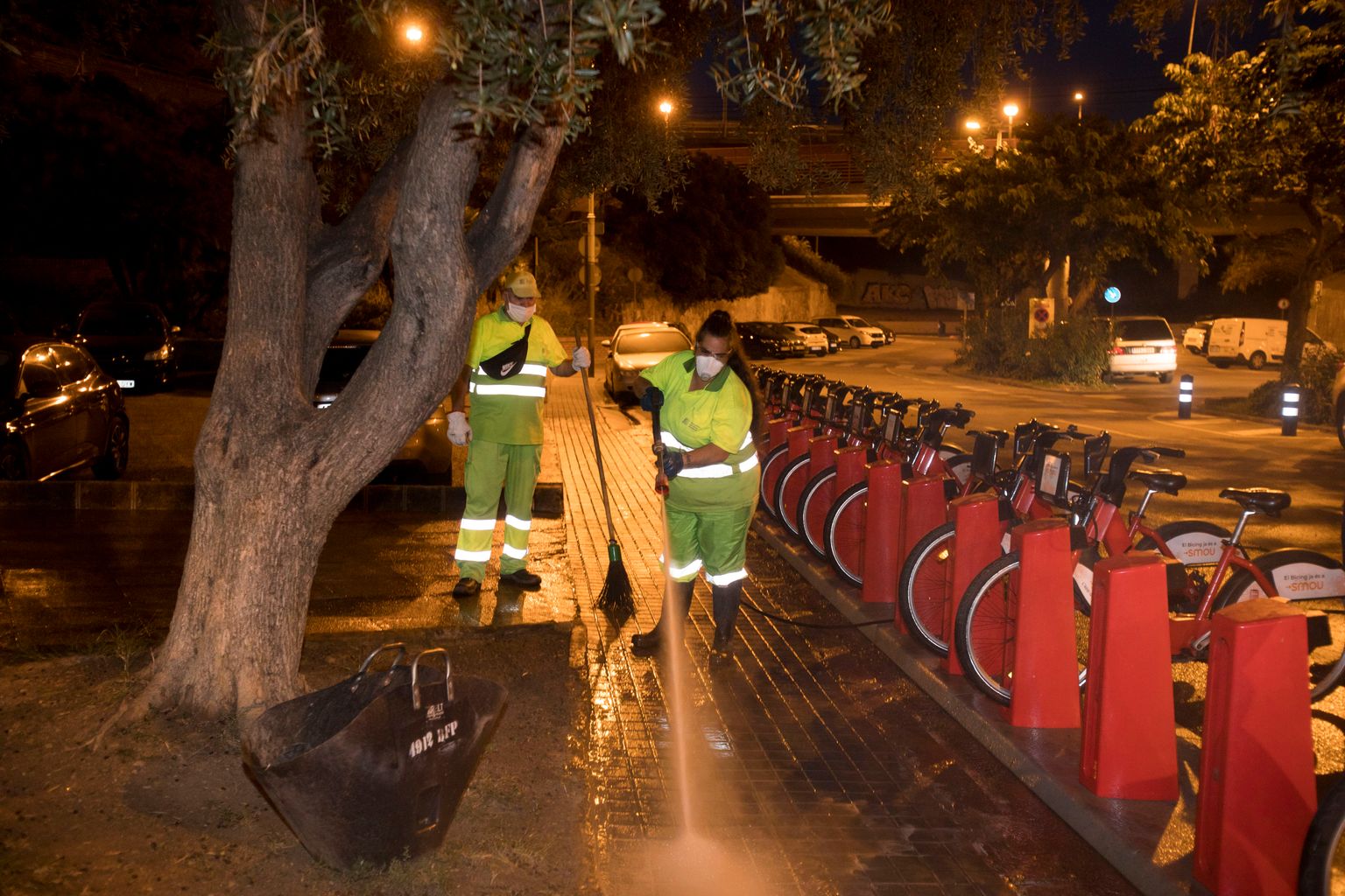 Una trabajadora del Servicio de Limpieza limpia con agua a presión la acera de alrededor de la estación de Bicing en la plaza Roja de la Ciutat Meridiana mientras un compañero espera