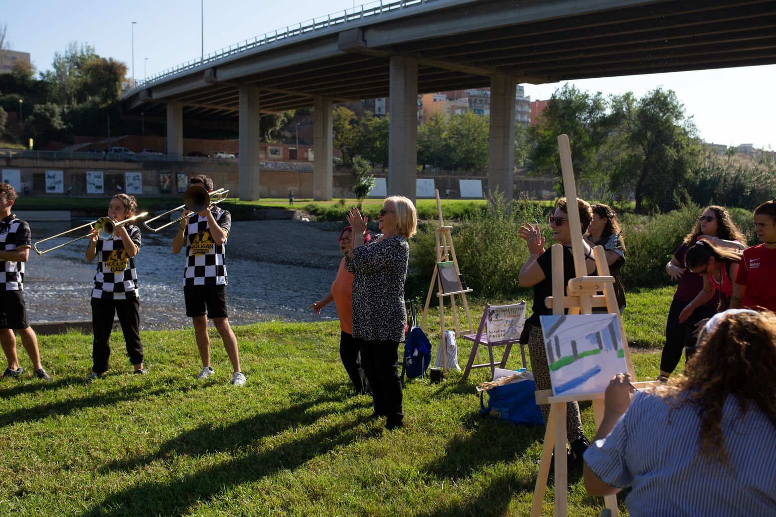 Un grup de gent pintant quadres a la gespa de la riba del riu Besòs anima un grup de nois tocant instruments de vent.
