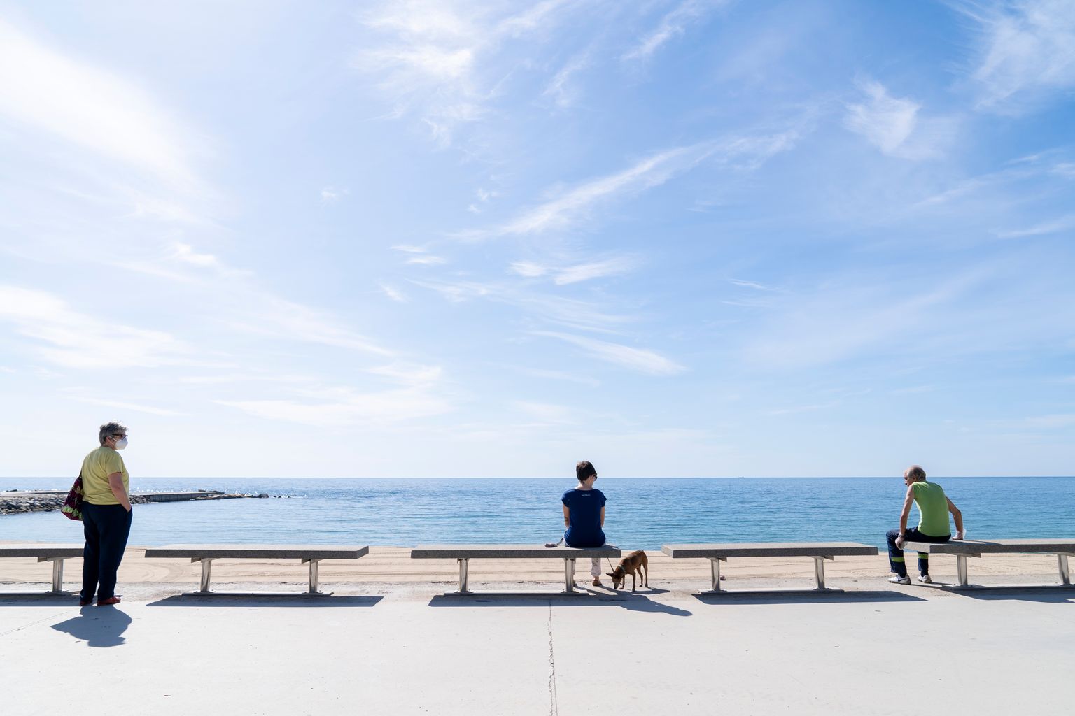 Persones assegudes als bancs del passeig Marítim del Bogatell contemplant el mar i guardant la distància de seguretat entre elles