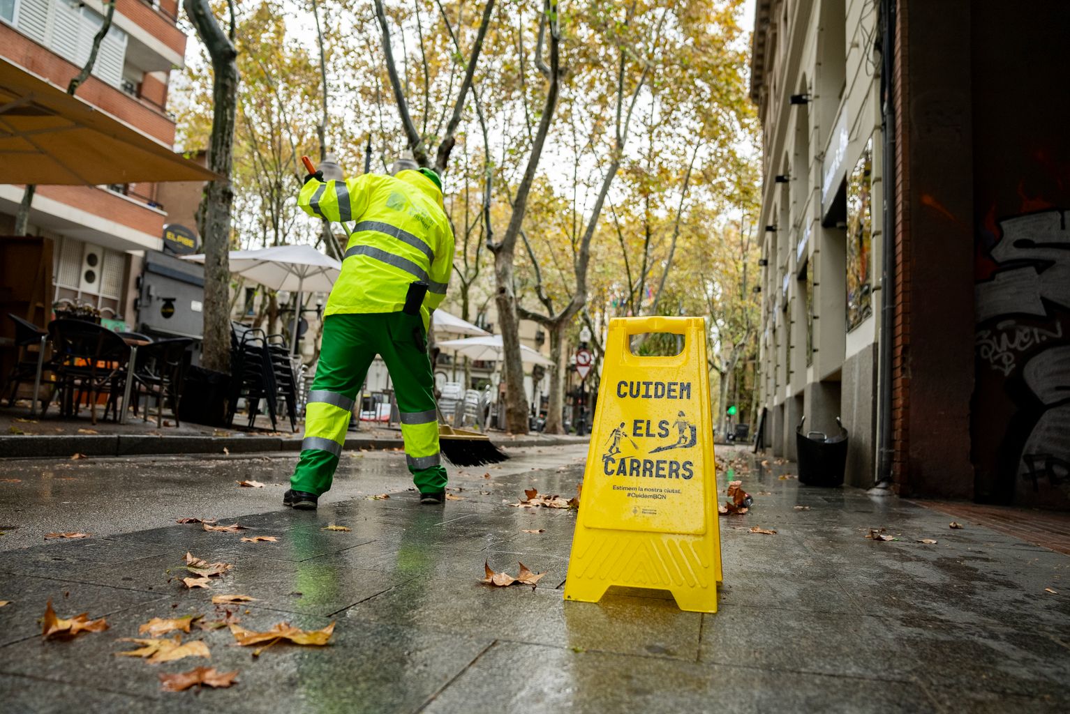 Un trabajador del Servicio de Limpieza barre las hojas secas de una acera de la rambla del Poblenou. Ha puesto un indicador de aviso con el letrero “Cuidem els carrers”