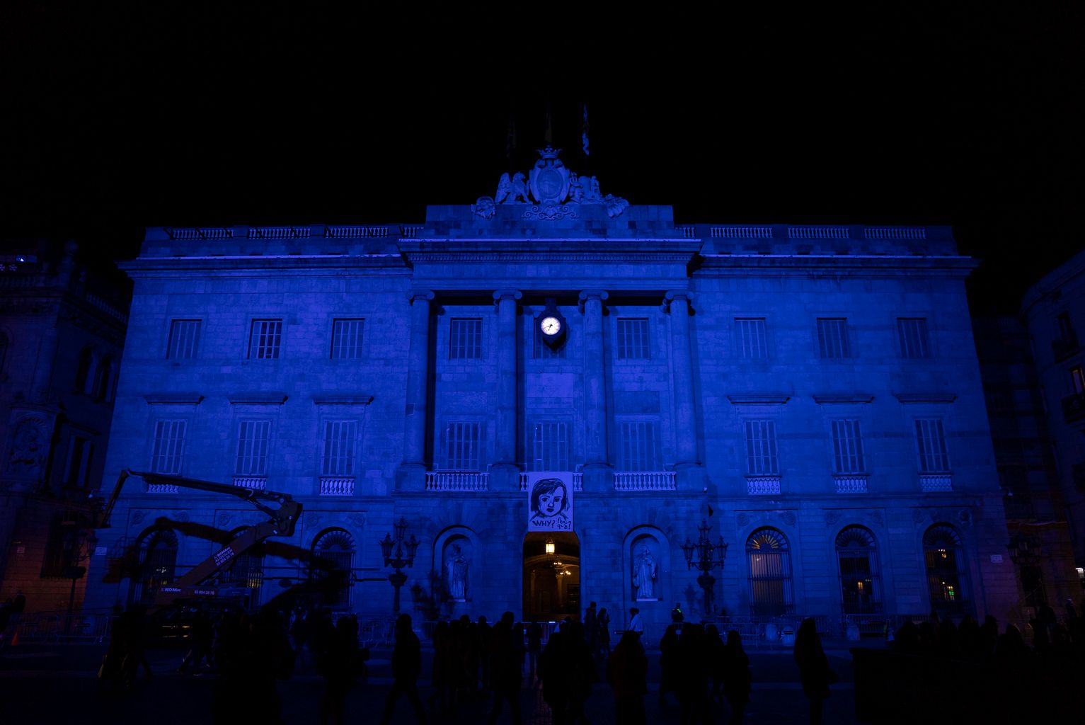 Façana de l'Ajuntament il·luminada de blau per commemorar el Dia Mundial de la Infància i la pancarta penjada al balcó amb gent a la plaça fent fotografies