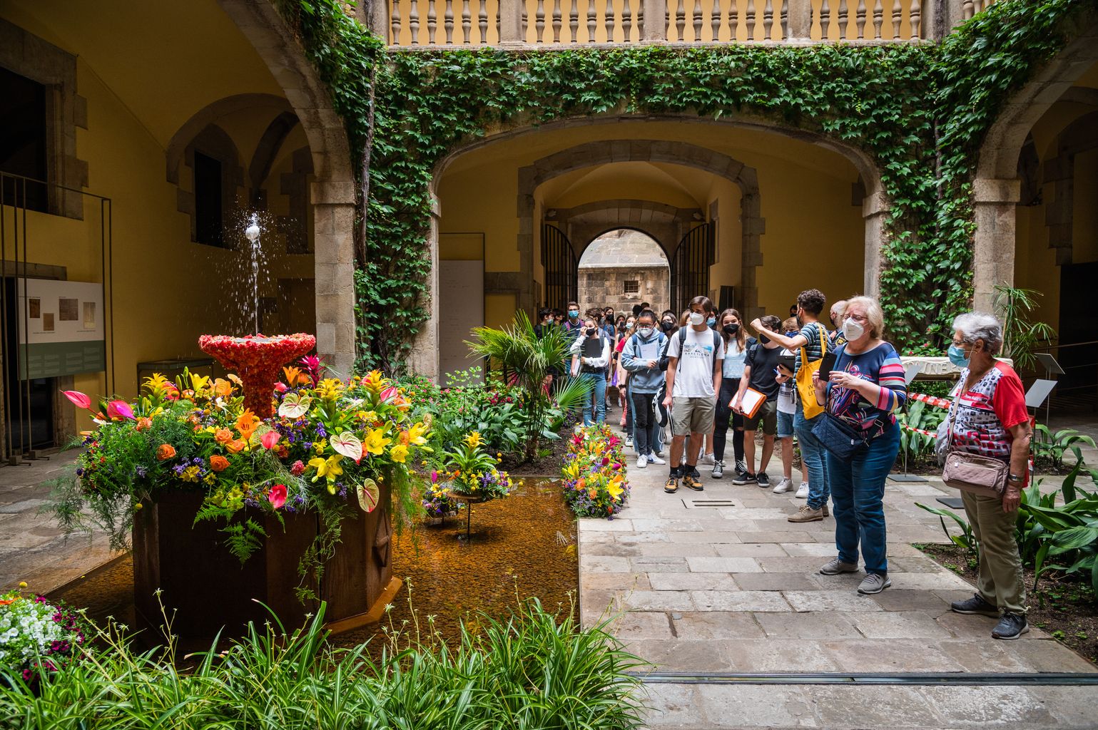 Gent fotografia l'ou com balla del pati del Palau del Lloctinent. El brollador està envoltat i guarnit amb flors