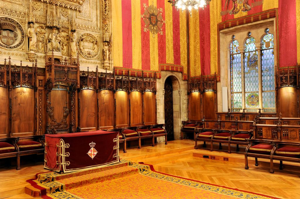 Saló de Cent decorat amb els tapissos de la senyera. Capçalera del saló amb el cadirat i retaule d'alabastre amb l'escut de Barcelona amb la sala