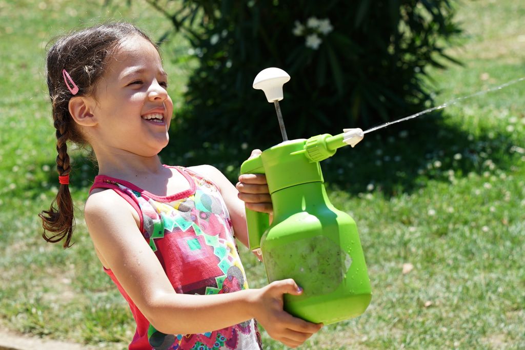 Una nena jugant amb un sifó que dispara aigua. La nena porta trenes, un vestit de flors i somriu.