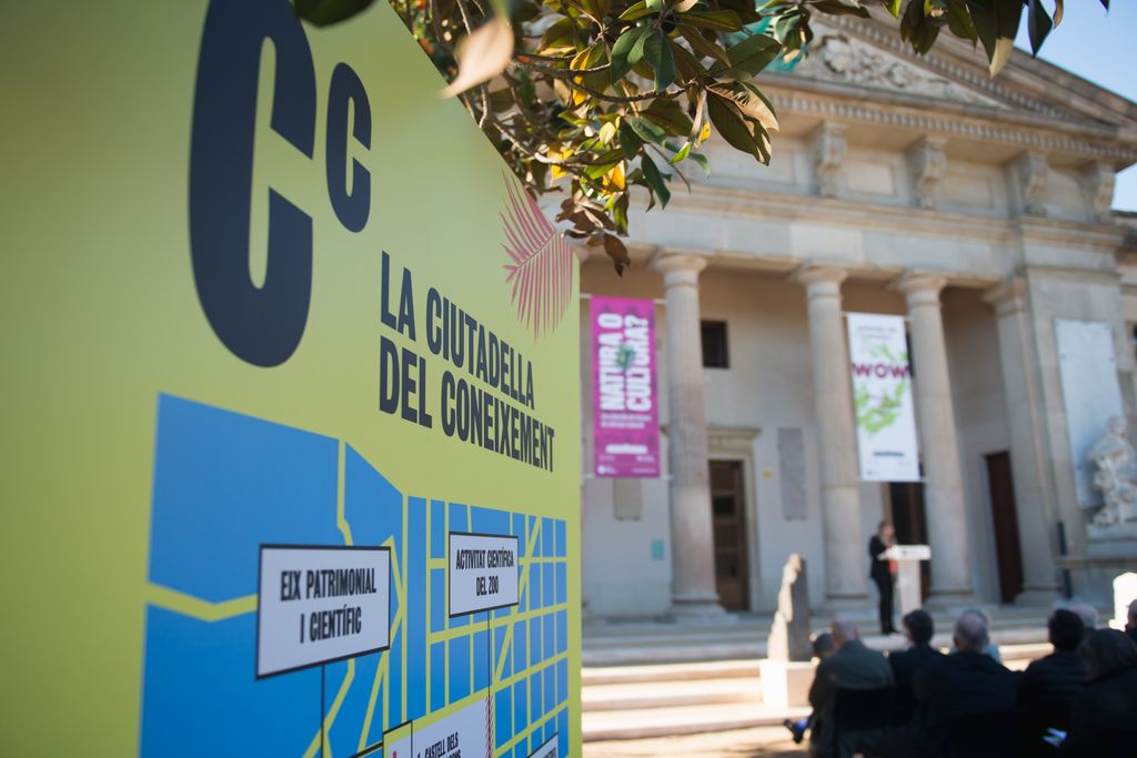 Cartell amb la imatge gràfica de la Ciutadella del Coneixement. De fons, l'acte d'inauguració del Centre Martorell d'Exposicions